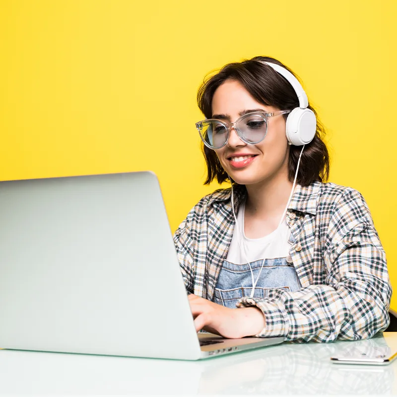 A smiling woman in headphones and glasses working on a laptop in front of a yellow wall